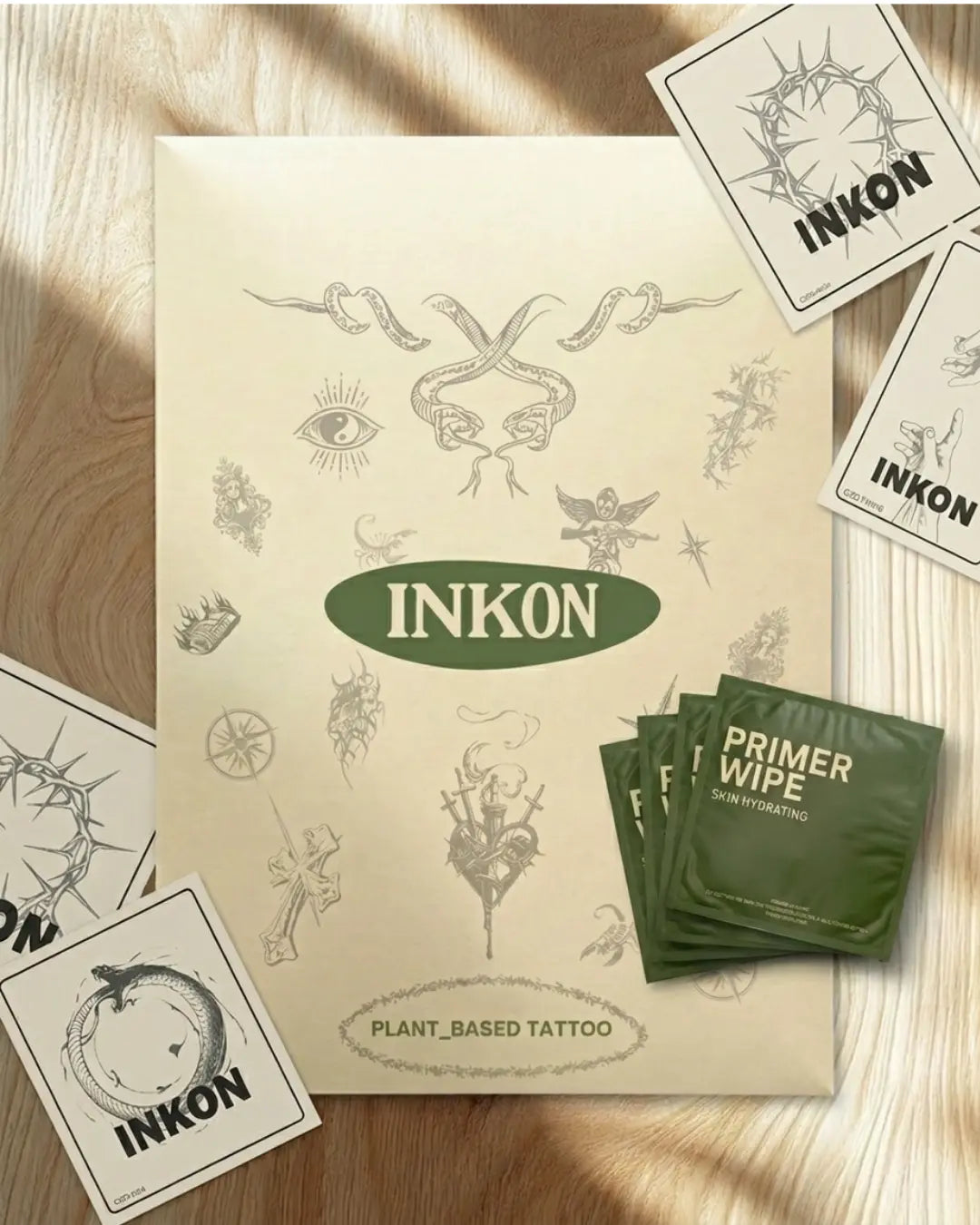 Kit di confezionamento completo INKON Tattoos For Now su superficie di marmo – busta verde brandizzata con illustrazioni floreali e di farfalle, salvietta primer idratante per la pelle, scheda con le istruzioni di applicazione con QR code e adesivo brandizzato INKON Tattoo